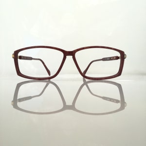 Könnte beinhalten: Eine Brille mit rechteckigen braunen Gläsern und goldenen Akzenten an den Bügeln. Die Brille spiegelt sich auf einer weißen Oberfläche.