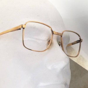 Op de afbeelding: Gouden metalen bril met vierkante glazen en zwarte pootjes. De bril zit op een wit mannequin hoofd.