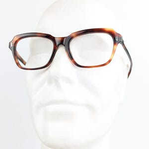 May include: Rectangular eyeglasses with a tortoiseshell pattern. The frames are brown and amber, with clear lenses. The glasses are displayed on a white mannequin head, showcasing their shape and design.