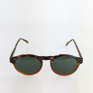 May include: Round sunglasses with a tortoiseshell frame and dark green lenses. The frames have a classic, retro design with a keyhole bridge. The arms are also tortoiseshell, providing a cohesive look. These sunglasses offer a stylish, timeless aesthetic.