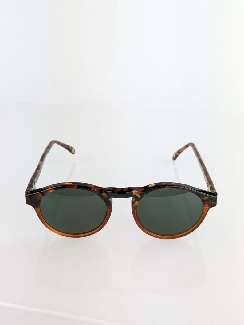 May include: Round sunglasses with a tortoiseshell frame and dark green lenses. The frames have a classic, retro design with a keyhole bridge. The arms are also tortoiseshell, providing a cohesive look. These sunglasses offer a stylish, timeless aesthetic.