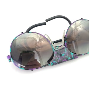 Peut inclure: Une paire de lunettes de soleil aviateur noires et violettes avec un motif floral sur la monture. Les verres sont bruns et les lunettes ont un design unique avec une charnière au milieu.
