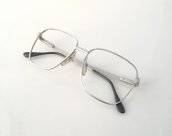 May include: Silver-framed eyeglasses with a rectangular shape. The glasses have clear lenses and black temples. The frame is made of a shiny metal, and the glasses are set against a white background.