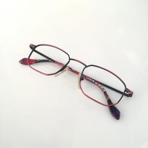 May include: Red and black rectangular eyeglasses with a floral pattern on the arms. The brand name "MIKLI" is printed on the right arm.