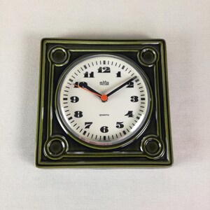 May include: A green ceramic wall clock with a white face and black numbers. The clock says "alfa hallen" and "quartz".