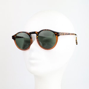 May include: Round sunglasses with a tortoiseshell pattern frame and dark green lenses. The sunglasses are displayed on a white mannequin head. The frame has a brown and black pattern.