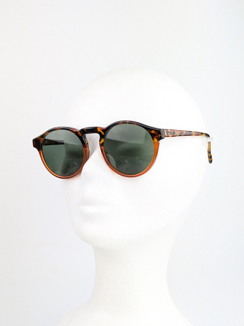 May include: Round sunglasses with a tortoiseshell pattern frame and dark green lenses. The sunglasses are displayed on a white mannequin head. The frame has a brown and black pattern.