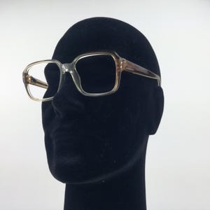 Könnte beinhalten: Brille mit transparentem Rahmen und rechteckiger Form. Die Rahmen gehen an den Bügeln in Braun über. Die Brille wird auf einem schwarzen Schaufensterkopf vor weißem Hintergrund präsentiert. Der Gesamtstil ist Vintage.