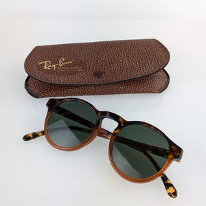 May include: Round tortoiseshell sunglasses with dark green lenses and a brown leather case. The sunglasses have a classic design with a keyhole bridge and are branded with the Ray-Ban logo.