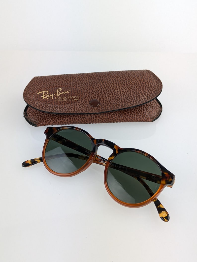 May include: Round tortoiseshell sunglasses with dark green lenses and a brown leather case. The sunglasses have a classic design with a keyhole bridge and are branded with the Ray-Ban logo.