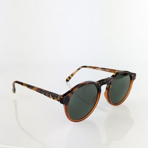 May include: Round sunglasses with a tortoiseshell frame and dark green lenses. The sunglasses have a classic design with a keyhole bridge and a subtle, polished finish. The arms of the sunglasses are also tortoiseshell.