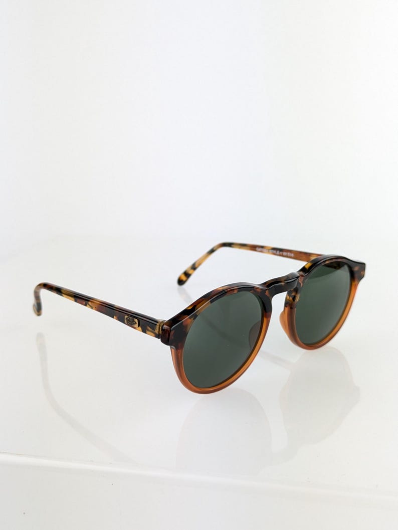 May include: Round sunglasses with a tortoiseshell frame and dark green lenses. The sunglasses have a classic design with a keyhole bridge and a subtle, polished finish. The arms of the sunglasses are also tortoiseshell.
