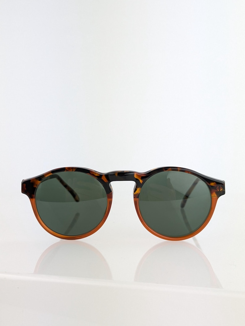May include: Round sunglasses with a tortoiseshell pattern frame and dark green lenses. The sunglasses have a classic design with a brown and black frame. The lenses are a dark green color, providing a stylish and functional accessory.