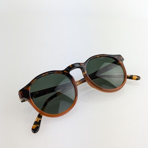 May include: Round sunglasses with a tortoiseshell pattern frame and dark green lenses. The sunglasses have a classic design with a keyhole bridge and are suitable for both men and women.