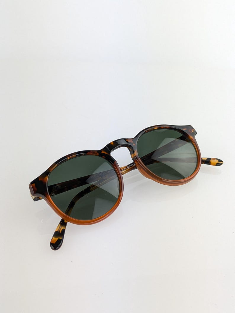 May include: Round sunglasses with a tortoiseshell pattern frame and dark green lenses. The sunglasses have a classic design with a keyhole bridge and are suitable for both men and women.
