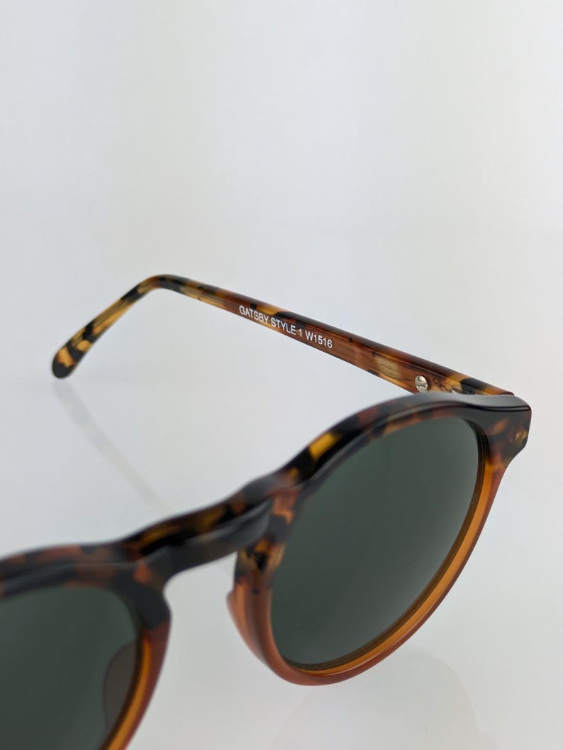 May include: Close-up of Gatsby style sunglasses with a tortoise shell pattern. The sunglasses have dark lenses and the text "Gatsby Style 1 W1516" is printed on the arm.