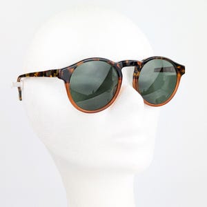 May include: Round sunglasses with a tortoiseshell frame and dark green lenses. The sunglasses have a classic design with a brown and orange rim. The sunglasses are displayed on a white mannequin head.