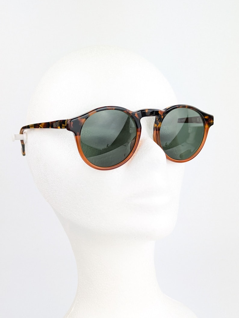 May include: Round sunglasses with a tortoiseshell frame and dark green lenses. The sunglasses have a classic design with a brown and orange rim. The sunglasses are displayed on a white mannequin head.