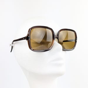 May include: Brown square-framed sunglasses with tinted lenses. The sunglasses have a dark brown frame and temples. The lenses are a light brown color. The sunglasses are displayed on a white mannequin head.