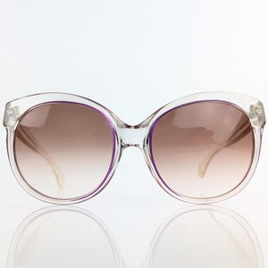 May include: Clear-framed sunglasses with a rounded shape and gradient brown lenses. The frames have a subtle purple accent around the lenses. The sunglasses are reflected on a white surface.
