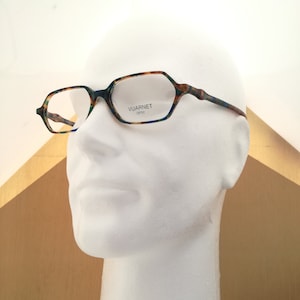 May include: A pair of tortoiseshell eyeglasses with a blue and green pattern. The glasses have a rectangular shape with rounded edges. The brand name "VUARNET OPTIC" is printed on the right lens.