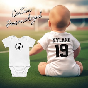 May include: White baby onesie with a black football graphic on the front and the text "NYLAND 19" on the back. The onesie is on a baby sitting on a green grassy field.