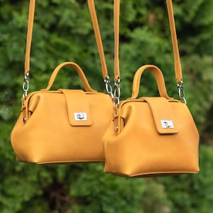 Puede incluir: Dos bolsos de mano de color amarillo mostaza con correas a juego cuelgan sobre un fondo verde borroso. Cada bolso tiene un asa superior, una solapa rectangular con un cierre plateado y una forma estructurada y cuadrada. Los bolsos son de tamaño similar.