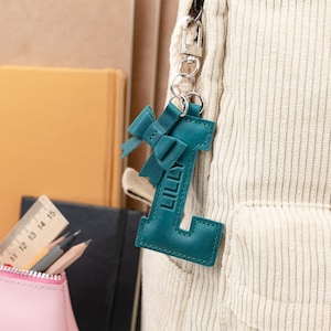 Op de afbeelding: Teal leren sleutelhanger in de vorm van de letter "L" met de naam "LILLY" erop gedrukt. De sleutelhanger heeft een strikdetail en is bevestigd aan een zilveren sluiting. De sleutelhanger hangt aan een corduroy rugzak.