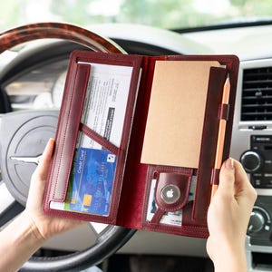 Op de afbeelding: Bordeaux leren documenthouder met meerdere vakken, met een notitieblok, potlood, creditcards en documenten. De houder is open, waardoor de inhoud zichtbaar is, en wordt voor het stuur van een auto gehouden.
