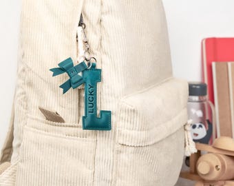 Peut inclure: Un sac à dos en velours côtelé crème avec un porte-clés en cuir bleu sarcelle. Le porte-clés a la forme de la lettre "L" avec le mot "LUCKY" et un nœud. Le sac à dos est sur une étagère blanche avec des livres et des jouets en bois.
