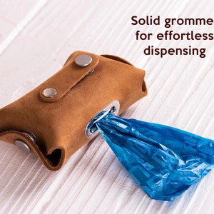 May include: Brown leather dog waste bag dispenser with a metal grommet and a blue plastic bag attached. The text "Solid grommet for effortless dispensing" is printed above the dispenser.