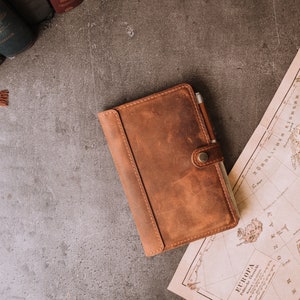 Pode incluir: Um di&aacute;rio de couro castanho com uma caneta presa por uma correia, sobre uma superf&iacute;cie cinza ao lado de um mapa vintage e livros. O di&aacute;rio tem um fecho de bot&atilde;o e uma apar&ecirc;ncia texturizada.