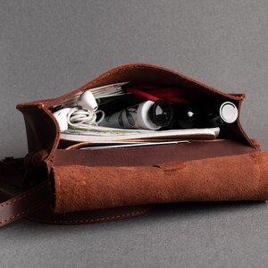 May include: A brown leather satchel bag with a flap closure. The bag is open and filled with various items, including a tube of hand cream, a pair of headphones, and a few magazines.