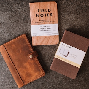 Pode incluir: Uma vista a&eacute;rea de tr&ecirc;s cadernos em uma superf&iacute;cie escura. Um caderno de madeira marrom claro com o texto "FIELD NOTES" est&aacute; no centro. Um caderno de couro marrom com fecho de bot&atilde;o est&aacute; &agrave; esquerda. Um caderno Moleskine marrom est&aacute; &agrave; direita.