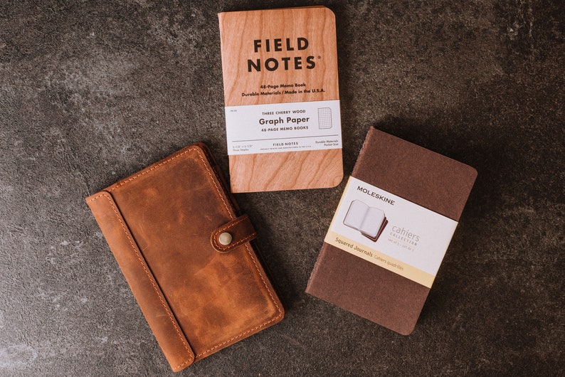 Pode incluir: Uma vista a&eacute;rea de tr&ecirc;s cadernos em uma superf&iacute;cie escura. Um caderno de madeira marrom claro com o texto "FIELD NOTES" est&aacute; no centro. Um caderno de couro marrom com fecho de bot&atilde;o est&aacute; &agrave; esquerda. Um caderno Moleskine marrom est&aacute; &agrave; direita.