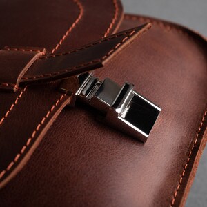 May include: Close-up of a brown leather bag with a silver metal clasp. The bag has a flap with a stitched edge and a strap.