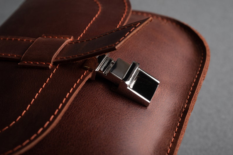 May include: Close-up of a brown leather bag with a silver metal clasp. The bag has a flap with a stitched edge and a strap.