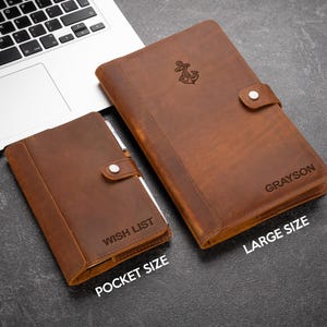 Pode incluir: Dois cadernos de couro castanho, um de bolso e outro grande, com fecho de bot&atilde;o. O caderno grande tem um emblema de &acirc;ncora e o nome "Grayson" gravado. O caderno de bolso diz "Wish List". Ambos est&atilde;o sobre uma superf&iacute;cie escura.