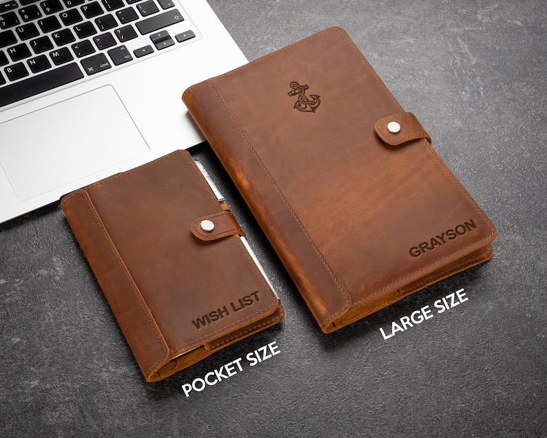 Pode incluir: Dois cadernos de couro castanho, um de bolso e outro grande, com fecho de bot&atilde;o. O caderno grande tem um emblema de &acirc;ncora e o nome "Grayson" gravado. O caderno de bolso diz "Wish List". Ambos est&atilde;o sobre uma superf&iacute;cie escura.