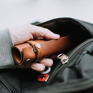 May include: A brown leather roll with a metal snap closure is being placed into a black zippered bag.