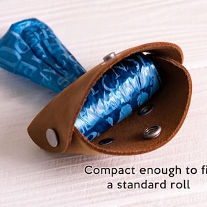 May include: A brown leather dog waste bag holder with a roll of blue plastic bags inside. The holder has a snap closure and metal rivets. Compact enough to fit a standard roll.