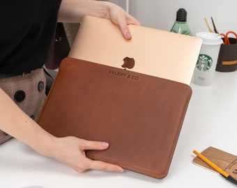 May include: A brown leather laptop sleeve with the text "VALERY & CO" embossed on the front. The sleeve is closed and a laptop is inside.