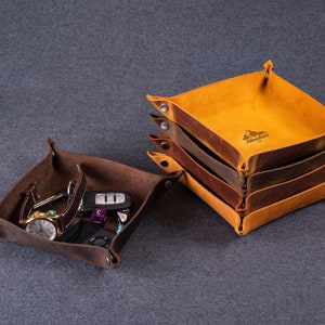 May include: Two leather trays, one brown and one yellow. The brown tray is filled with various small items, including a watch, keys, and a phone. The yellow tray is stacked on top of three other trays, all of which are brown. The trays are made of leather and have a distressed look.