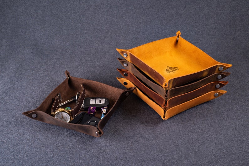 May include: Two leather trays, one brown and one yellow. The brown tray is filled with various small items, including a watch, keys, and a phone. The yellow tray is stacked on top of three other trays, all of which are brown. The trays are made of leather and have a distressed look.