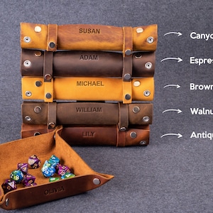 May include: A set of six leather dice trays in various shades of brown, including canyon, espresso, brown caramel, walnut, and antique cherry. Each tray is personalized with a name and features a snap closure. The trays are stacked on top of each other, with the top tray showing the name "SUSAN". A smaller leather dice tray with the name "OLIVIA" is in the foreground, filled with colorful dice.