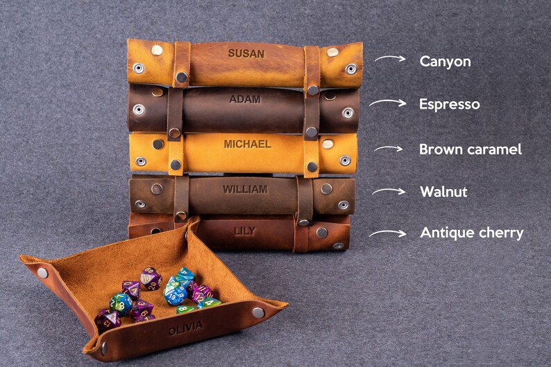 May include: A set of six leather dice trays in various shades of brown, including canyon, espresso, brown caramel, walnut, and antique cherry. Each tray is personalized with a name and features a snap closure. The trays are stacked on top of each other, with the top tray showing the name "SUSAN". A smaller leather dice tray with the name "OLIVIA" is in the foreground, filled with colorful dice.