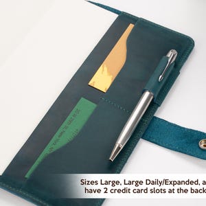 Pode incluir: Um di&aacute;rio de couro verde-azulado com uma caneta, um cart&atilde;o dourado e um cart&atilde;o verde nos bolsos. O di&aacute;rio est&aacute; aberto, revelando p&aacute;ginas brancas em branco. O texto na imagem diz: "Sizes Large, Large Daily/Expanded, and A5 have 2 credit card slots at the back side."