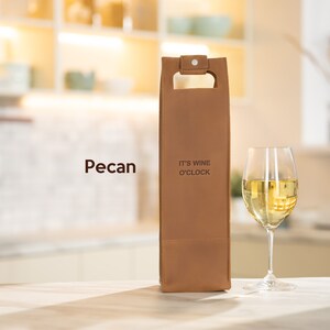 Pecan tan engraved leather wine bottle holder with custom text, romantic wine gift idea for husband or partner shown with white wine glass. A brown leather wine bottle carrier with the text "It's Wine O'Clock" printed on the front.