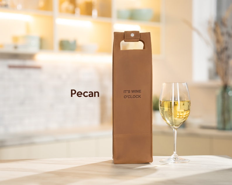 Pecan tan engraved leather wine bottle holder with custom text, romantic wine gift idea for husband or partner shown with white wine glass. A brown leather wine bottle carrier with the text "It's Wine O'Clock" printed on the front.