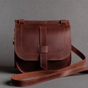 May include: A brown leather crossbody bag with a flap closure and an adjustable strap. The bag has a stitched design and a simple, classic style.